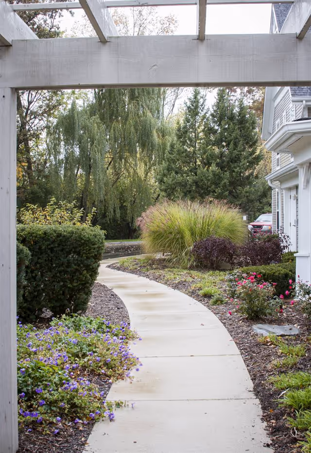 Curved concrete walkway through a landscaped garden beside a white residential building, viewed under a pergola.