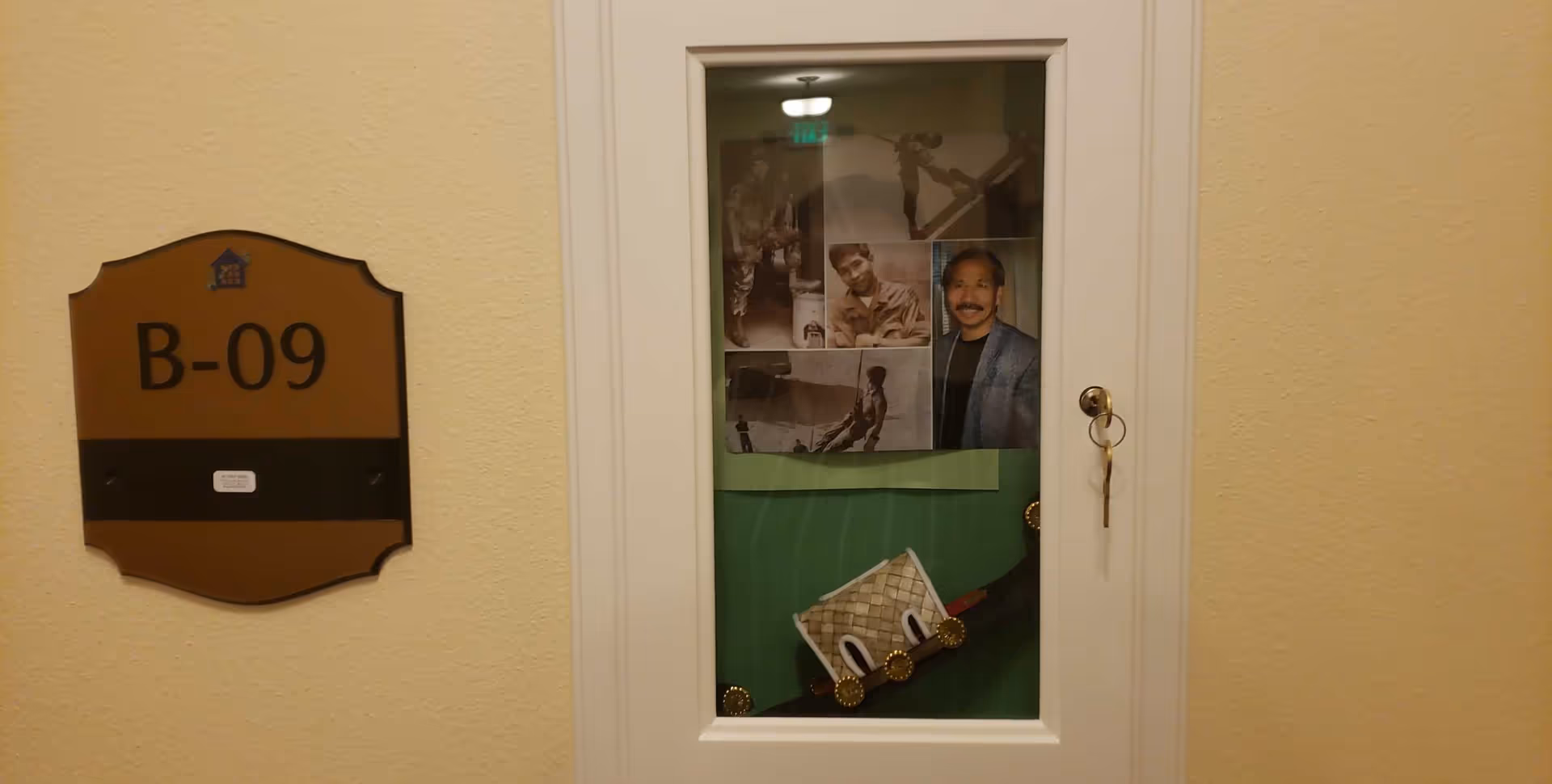 Hallway door marked B-09 with a glass inset displaying photos and a key hanging from the knob on a beige wall.