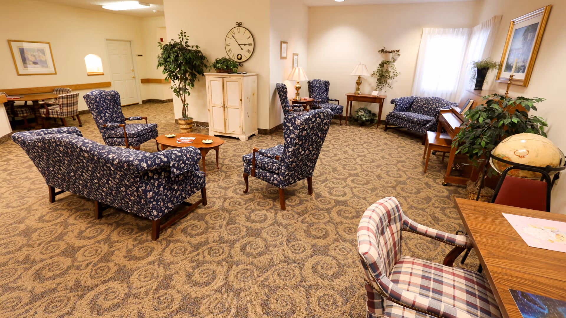 A cozy living room area with patterned blue armchairs and sofas arranged around a wooden coffee table. The room features a patterned carpet, a large wall clock, potted plants, a piano, framed artwork, and a table with a plaid chair in the foreground. Soft lighting and a window with sheer curtains brighten the space.