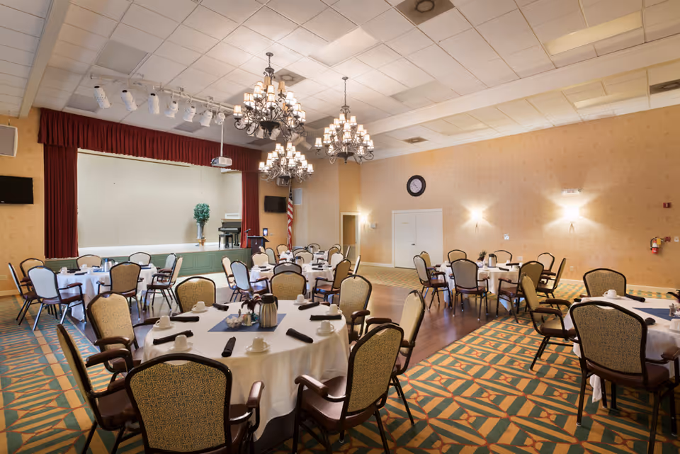 A spacious dining room with round tables covered in white tablecloths, each set with cups, saucers, and napkins. The room features patterned carpet, upholstered chairs, chandeliers hanging from the ceiling, and a small stage with a piano and an American flag in the background.