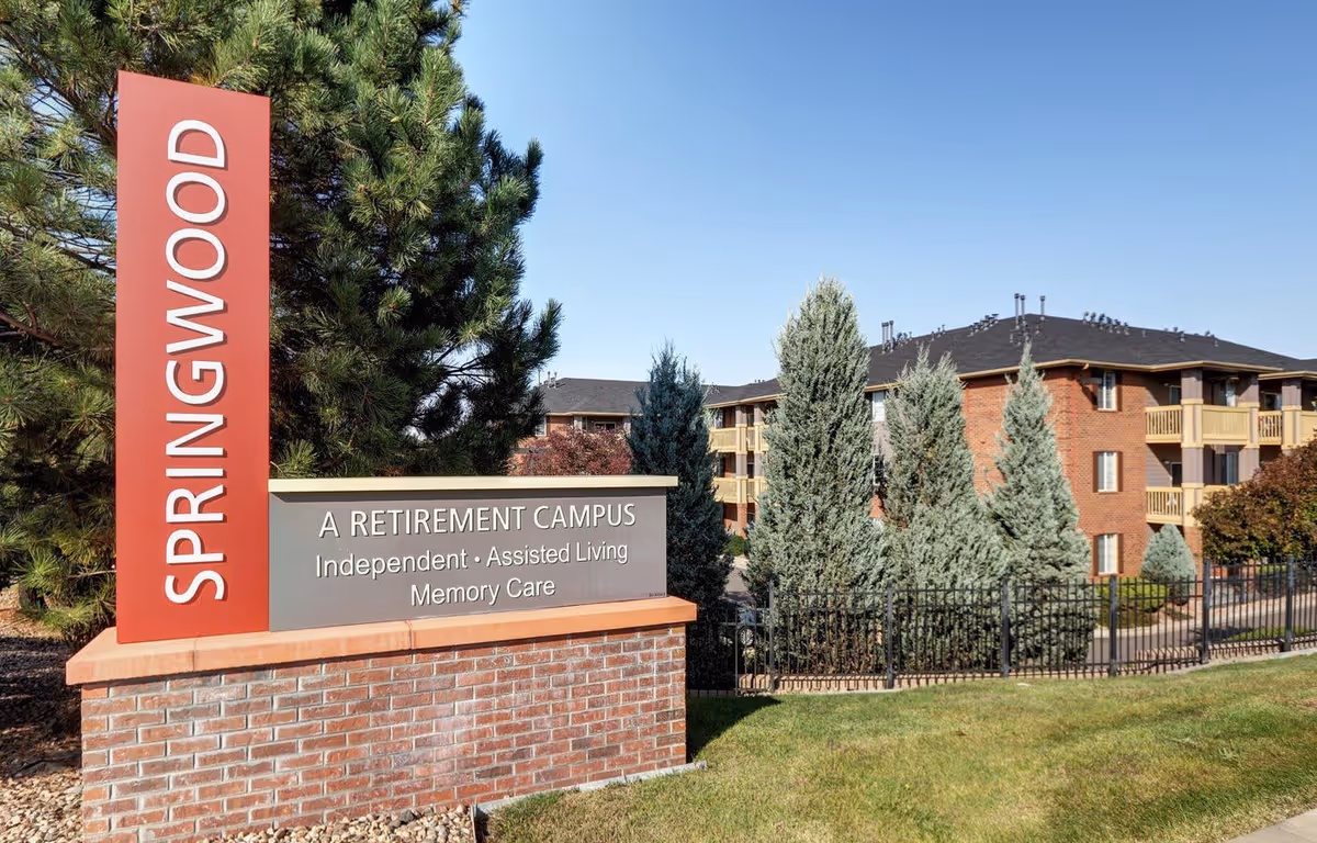 Outdoor view of the Springwood retirement campus sign with a brick base and red vertical panel, surrounded by trees and greenery, with a multi-story brick building in the background under a clear blue sky.