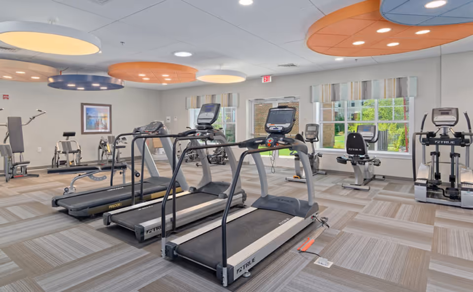 A bright and spacious fitness room with multiple exercise machines including treadmills, stationary bikes, and elliptical trainers. The room has large windows letting in natural light, modern ceiling lights with circular orange and white panels, and a patterned carpeted floor.