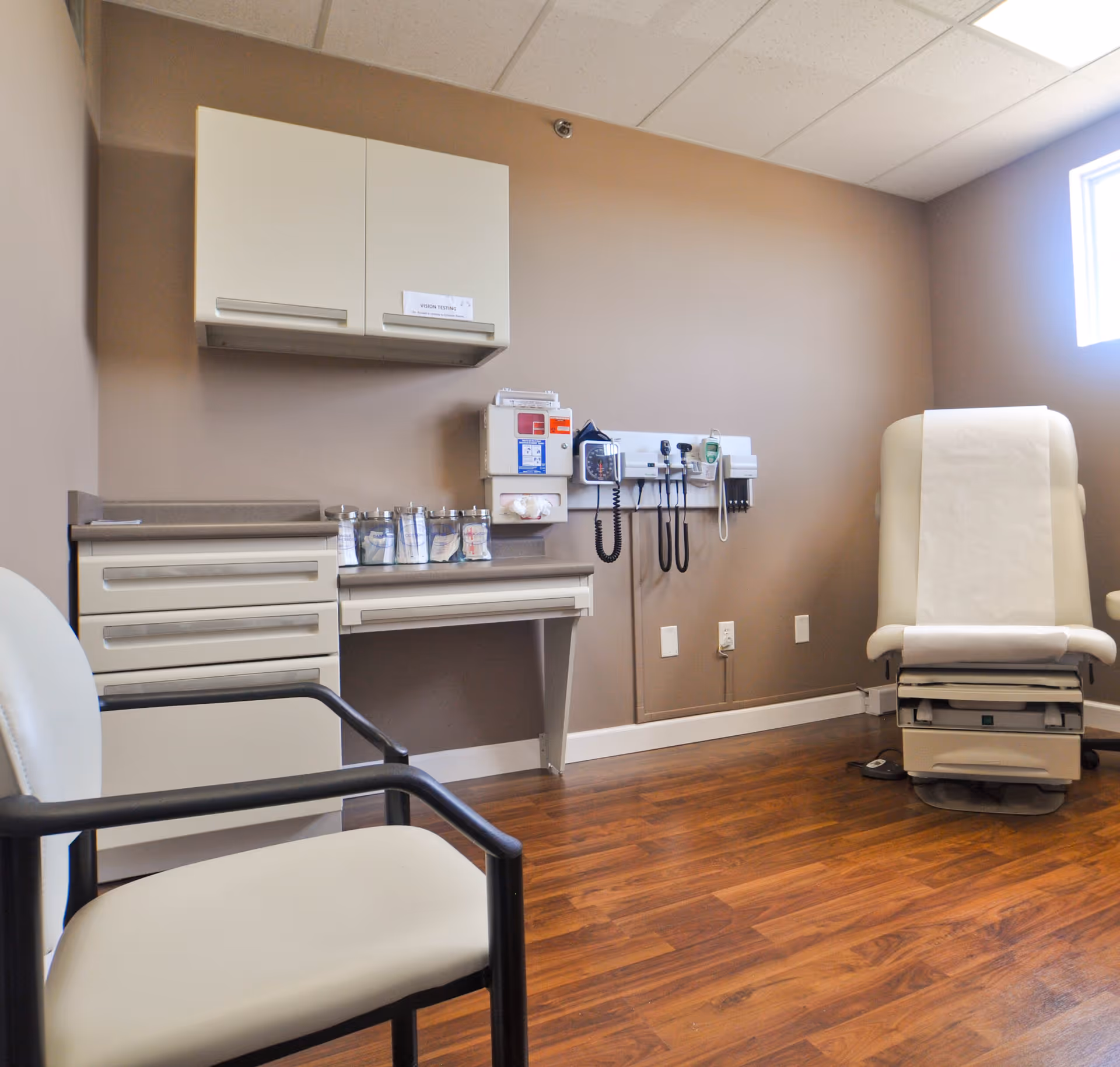 A medical examination room with a beige examination chair covered with white paper, a white chair with black armrests, a countertop with medical supplies, cabinets, and medical instruments mounted on the wall. The room has wood flooring and beige walls with a small window letting in natural light.