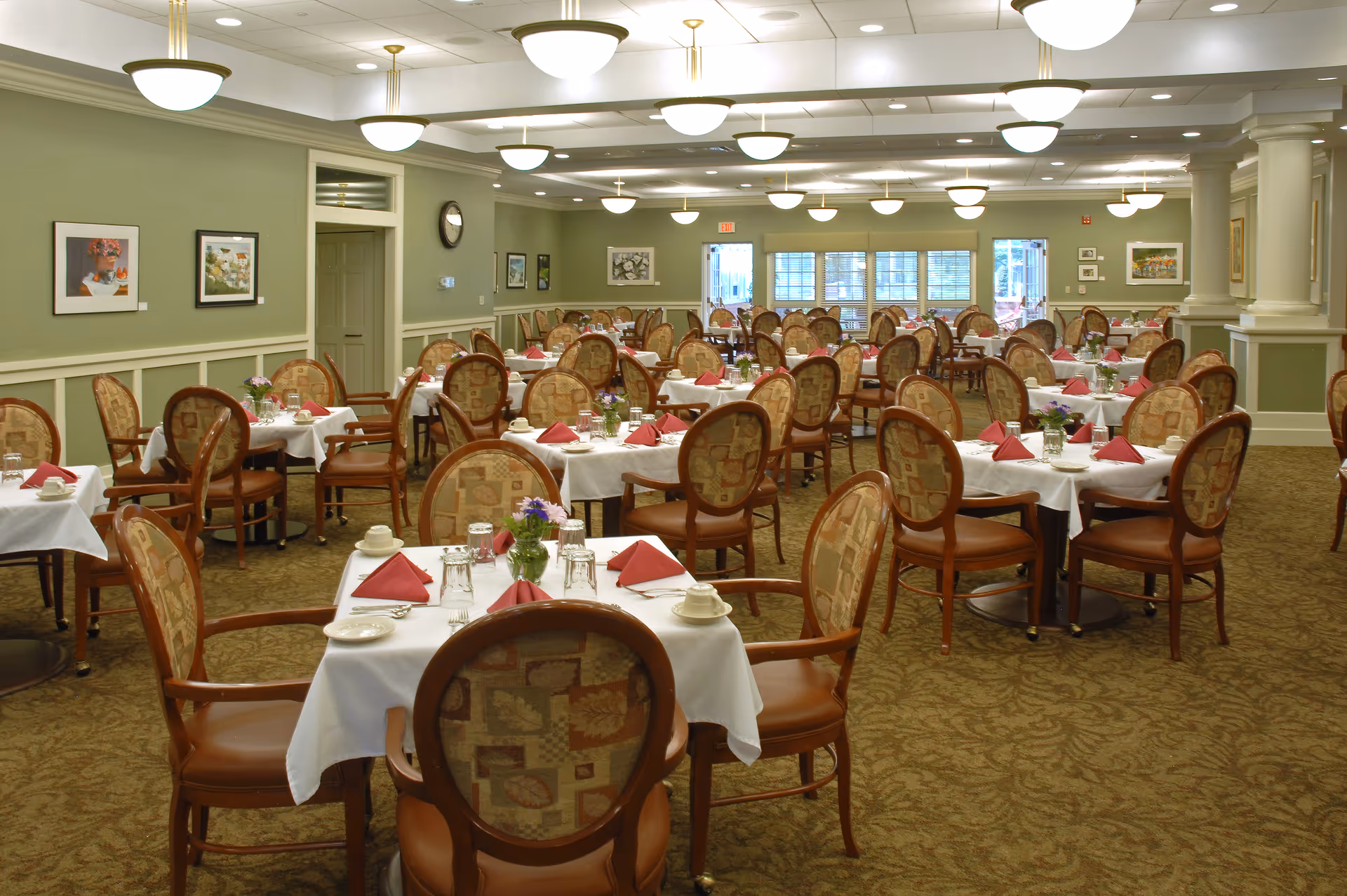 A spacious senior living dining room with multiple round tables covered with white tablecloths, each set with red folded napkins, glassware, cups, and small flower arrangements. The room has patterned carpet, green walls with framed artwork, and ceiling lights evenly spaced across the ceiling.