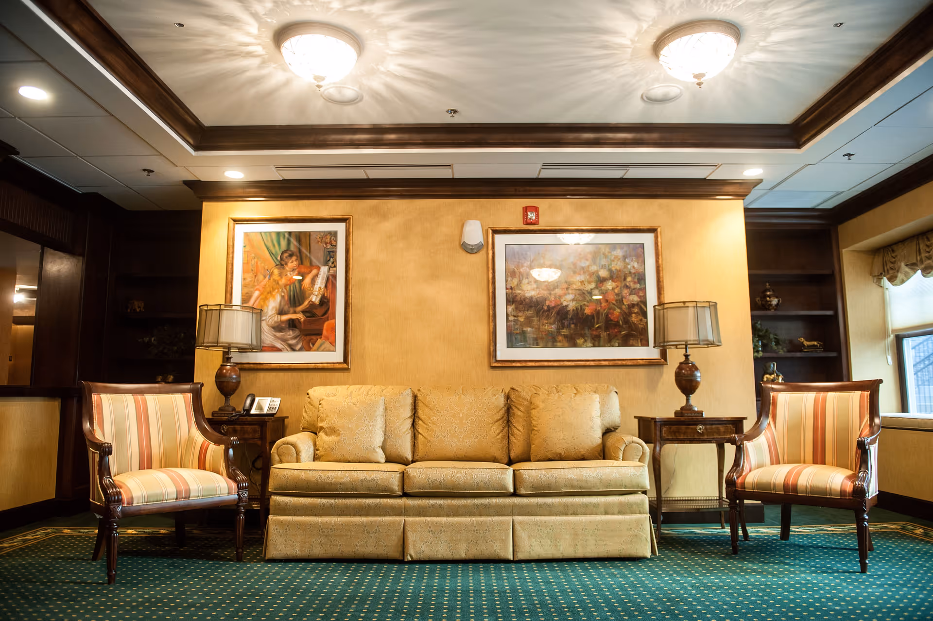 A cozy living room area with a beige sofa and two striped armchairs on either side. There are two wooden side tables with lamps on them, and two framed paintings hanging on a yellow wall behind the sofa. The room has a green carpet with a dotted pattern and a ceiling with recessed lighting and decorative light fixtures.