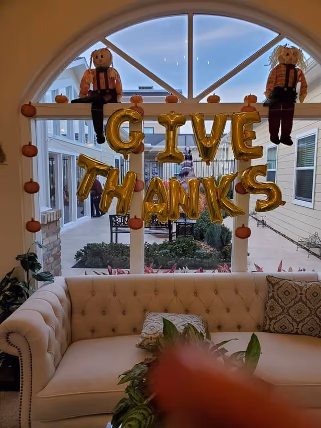Tufted sofa in a communal living area beneath a window decorated with gold 'GIVE THANKS' balloons and fall decorations.