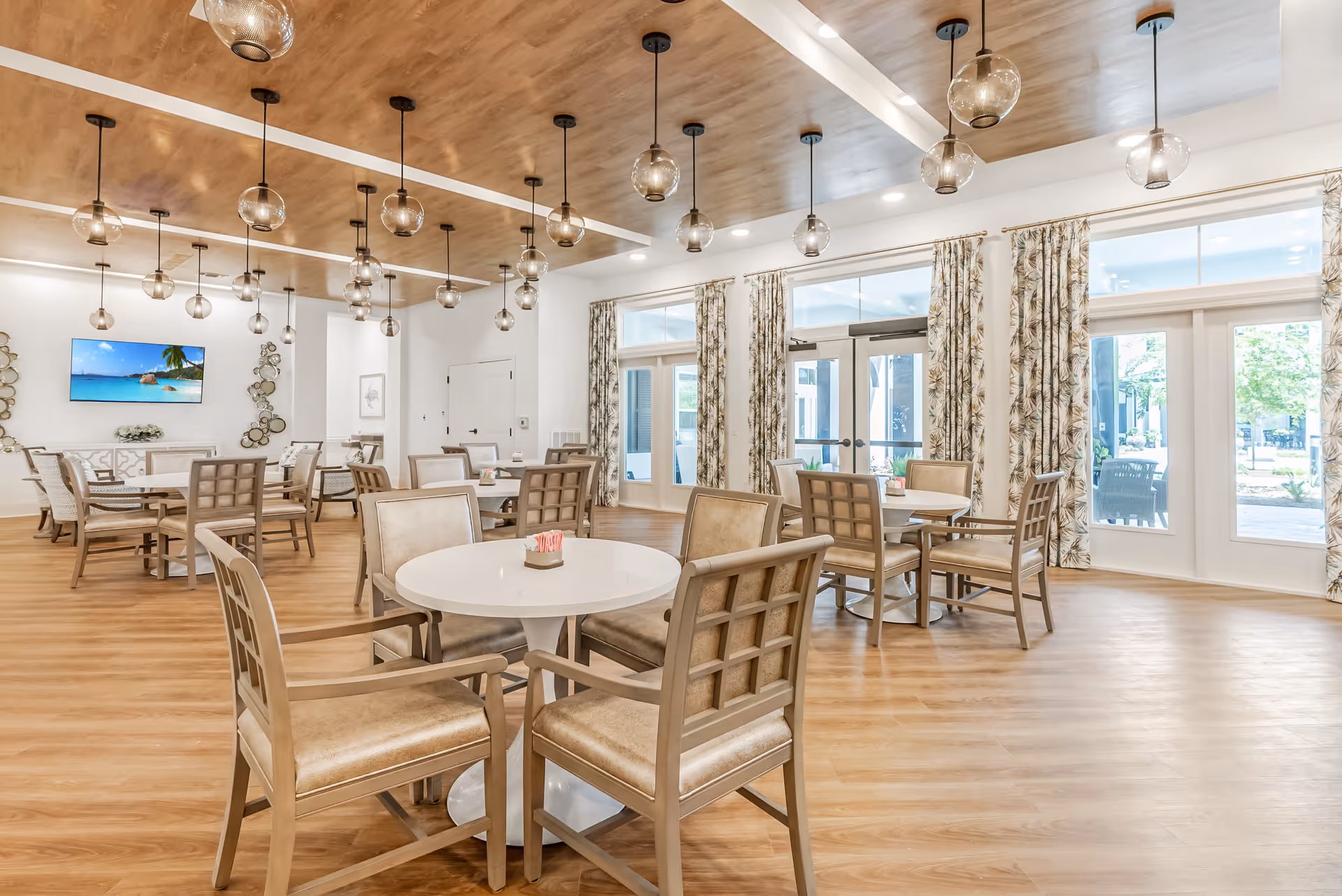 A bright and spacious dining room with multiple round tables and cushioned chairs. The room features large windows with patterned curtains allowing natural light to fill the space. The ceiling has wooden paneling with multiple hanging globe pendant lights. A flat-screen TV is mounted on the far wall, and the floor is covered with light wood flooring.