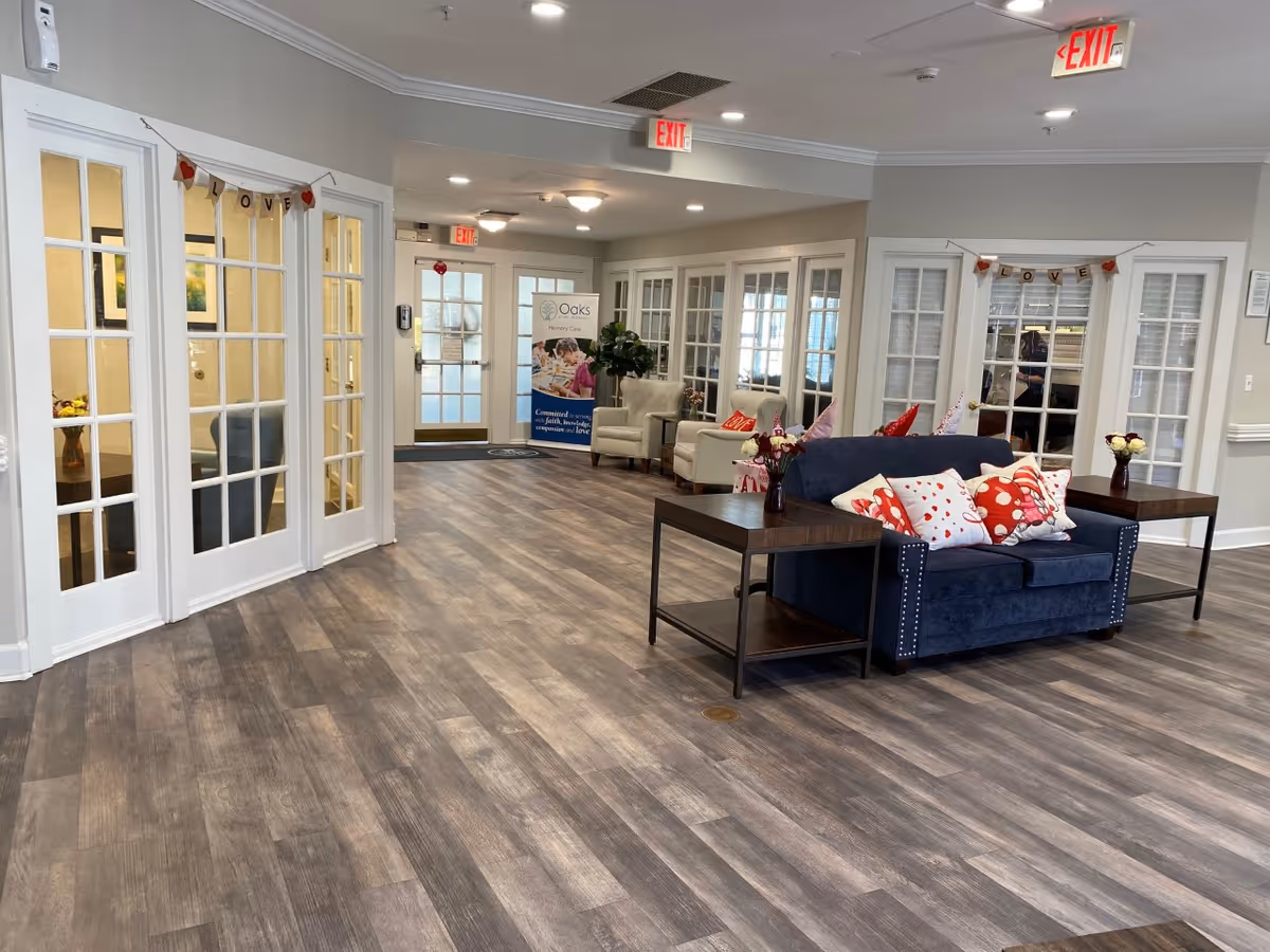 A bright and spacious common area in a senior living facility with wood-look flooring, a navy blue loveseat adorned with red and white heart-themed pillows, two side tables with flower vases, and beige armchairs in the background. The walls are light gray with white trim, and there are multiple glass-paneled doors. A banner with the word 'LOVE' and red hearts hangs above two sets of doors. Exit signs are visible on the ceiling, and a sign for Oaks Memory Care is seen near the back door.