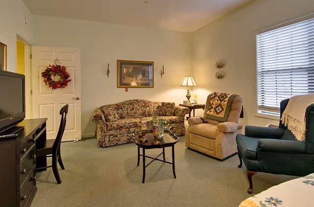 Cozy living room with a floral sofa, two armchairs, a coffee table, TV on a dresser, and a lamp by a window.