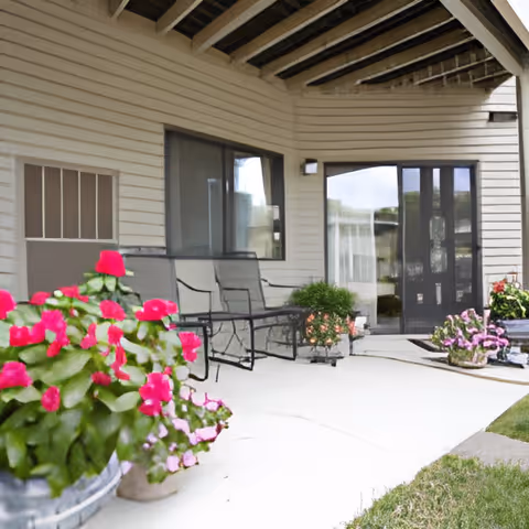 Covered outdoor patio area with metal chairs and tables, surrounded by potted plants with pink and red flowers. The patio is adjacent to a building with beige siding and large windows and doors.