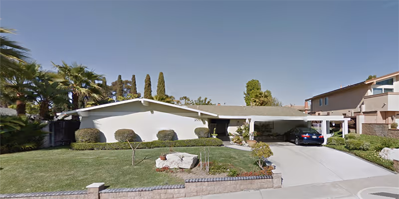 Front exterior view of a single-story residential building with a well-maintained lawn, shrubs, and a driveway with a parked car under a carport. The sky is clear and blue.