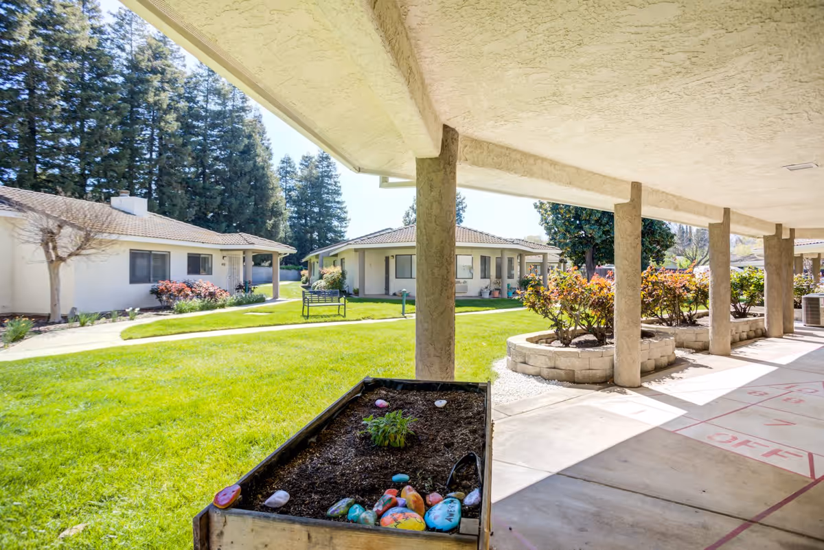 View of a sunny outdoor courtyard area at a senior living facility with green grass, small bushes, and several single-story buildings with tiled roofs. There is a covered walkway with columns and a raised garden bed containing soil and colorful painted rocks.