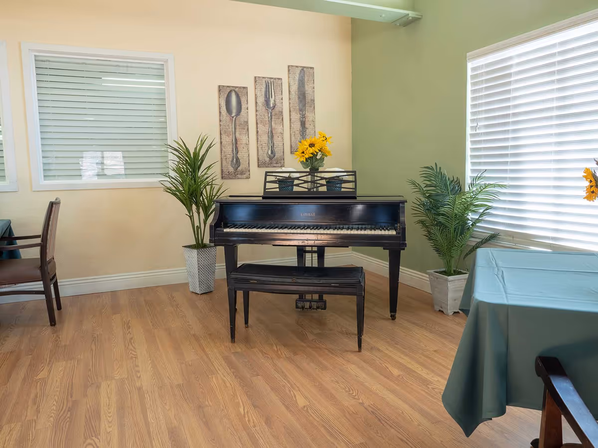 Interior room with a black upright piano and matching bench placed against a corner wall. The wall behind the piano is painted green and beige, decorated with three vertical art pieces depicting a spoon, fork, and knife. There are two potted plants on either side of the piano and a vase with yellow flowers on top. The room has wooden flooring, a window with white blinds on the right, and tables with green tablecloths and chairs partially visible on the left and right edges.