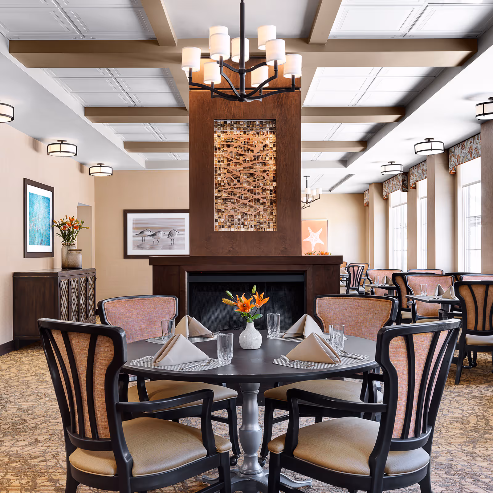 A well-lit dining room in a senior living facility featuring a round table set for four with beige napkins, glassware, and a small vase of orange lilies. The room has a central fireplace with decorative tile and wood paneling, beige walls, framed artwork, and multiple windows with patterned valances allowing natural light. Additional dining tables and chairs are visible in the background.