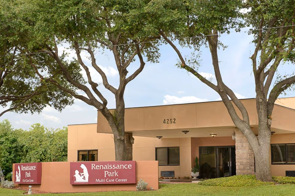 Exterior view of Renaissance Park Multi Care Center building with beige walls, large windows, and a covered entrance. Two large trees are in front of the building, and a sign with the facility name is visible near the entrance.