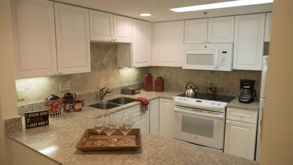 A modern kitchen with white cabinets and granite countertops. The kitchen features a double sink, an electric stove with an oven, a microwave, a coffee maker, and a refrigerator. There are decorative red canisters and a small sign that reads 'I KNOW HOW TO COOK. I JUST DON'T'. A wooden tray with four empty wine glasses is placed on the countertop.