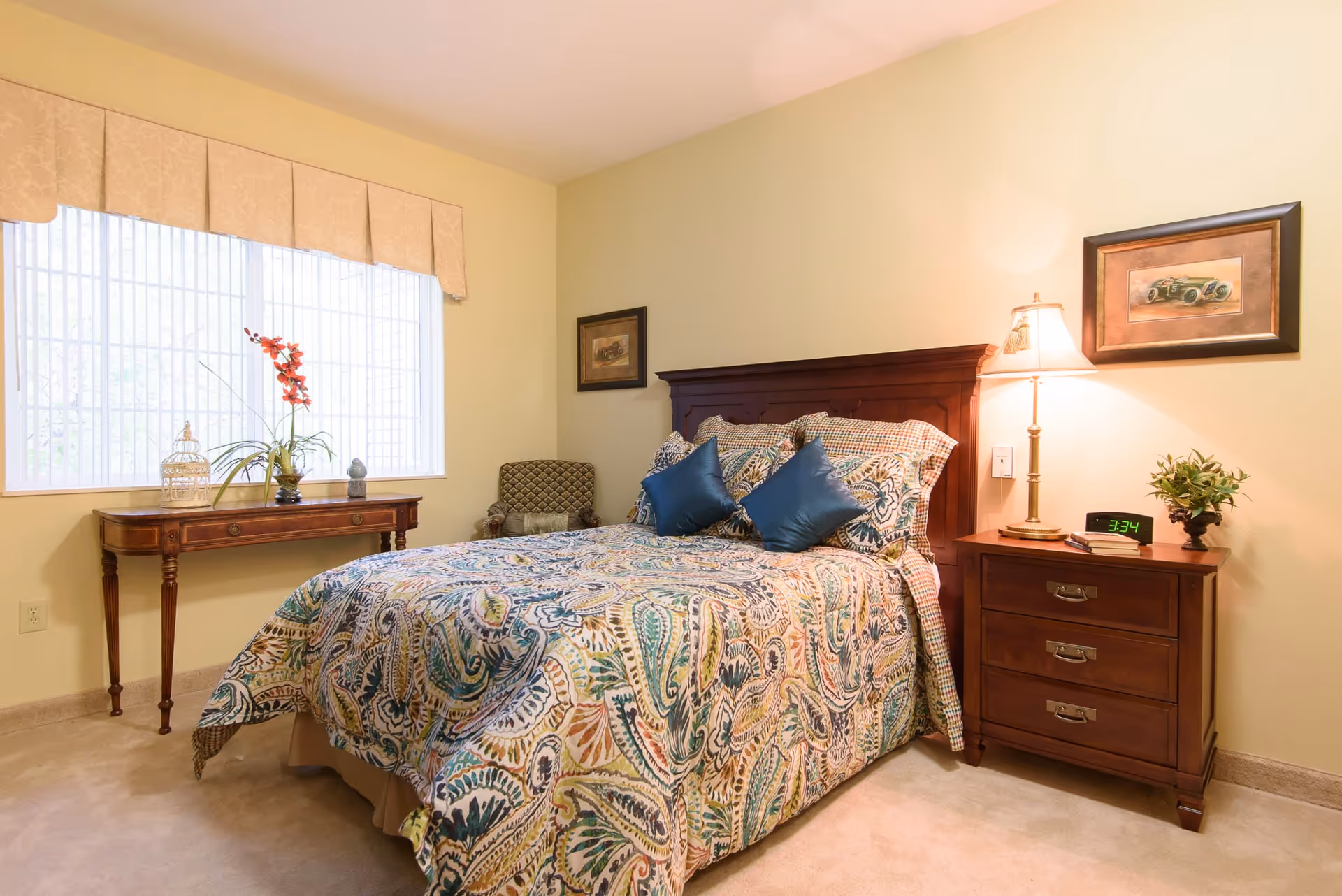 A cozy bedroom with a large window covered by a beige valance. The bed has a wooden headboard and is dressed with a colorful paisley-patterned bedspread and two blue accent pillows. To the right of the bed is a wooden nightstand with a lamp, a digital clock showing 3:34, a small plant, and a couple of books. On the left side of the room, there is a wooden console table with decorative items including a birdcage, a vase with red flowers, and a small sculpture. A patterned armchair is placed in the corner between the bed and the console table. The walls are painted a soft yellow color.