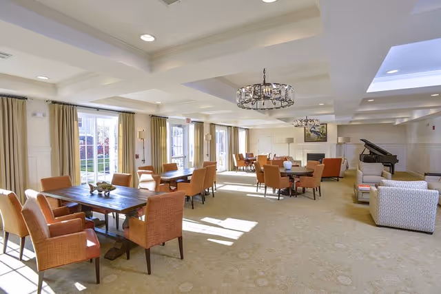 A spacious, well-lit common area with multiple wooden tables surrounded by orange upholstered chairs. The room features large windows with beige curtains allowing natural light to fill the space. There are chandeliers hanging from the ceiling, a fireplace with a painting above it, and a grand piano in the corner. Comfortable armchairs are arranged near the piano, creating a cozy seating area.