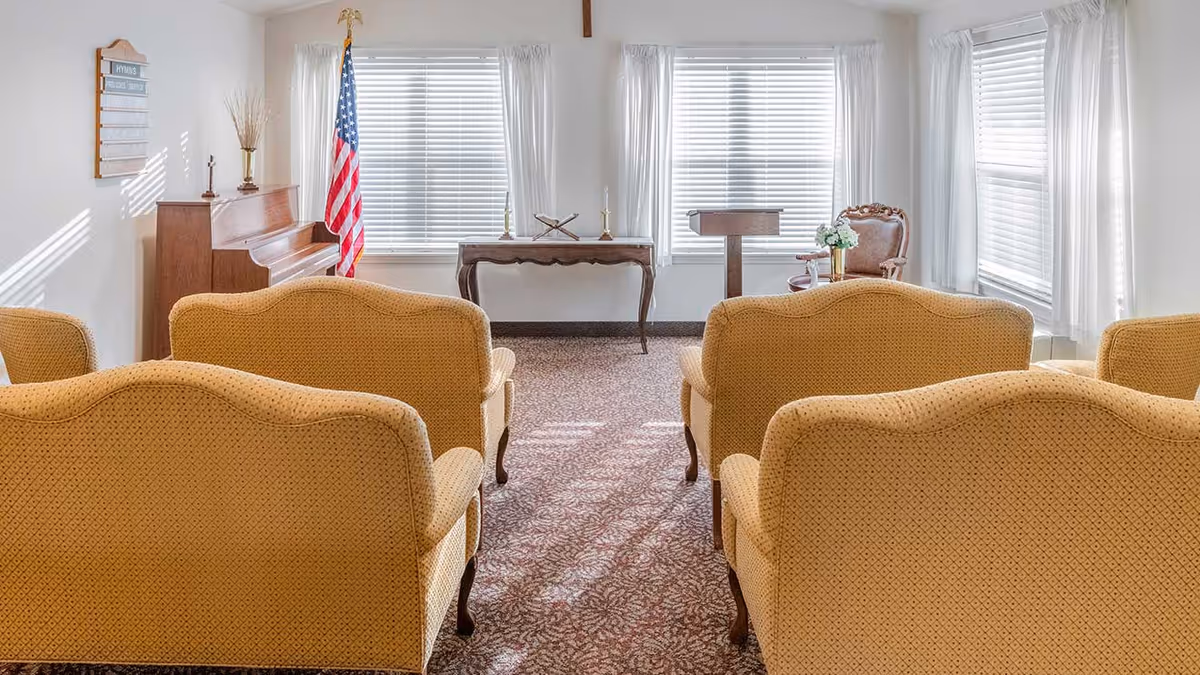 A small chapel or meeting room with rows of yellow upholstered chairs facing a wooden piano, an American flag, a small table with candlesticks, and a lectern. The room has large windows with white blinds and sheer white curtains, and a carpeted floor with a floral pattern.