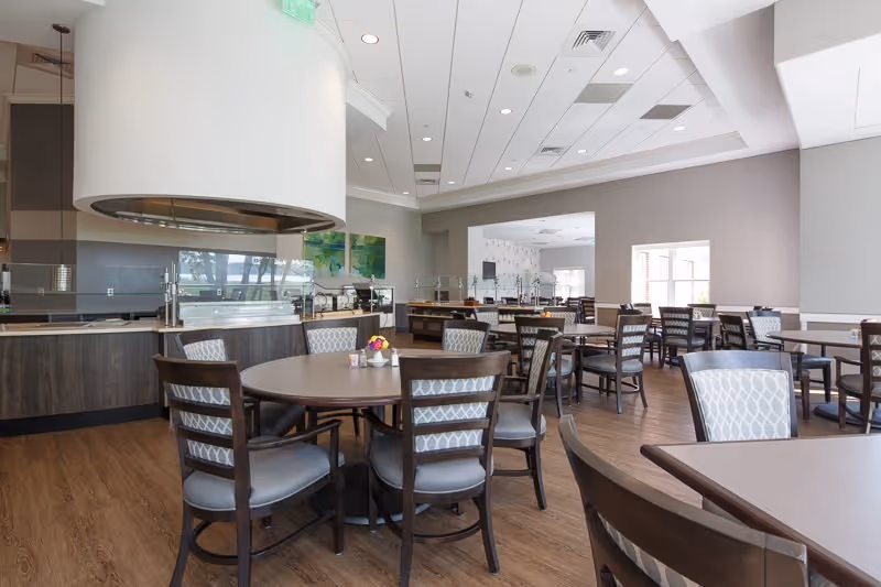 A spacious dining room with multiple round and rectangular tables surrounded by chairs. The room features wood flooring, neutral-colored walls, and a modern ceiling with recessed lighting. There is a serving area with a curved overhead vent and glass partitions, and large windows allowing natural light to enter.