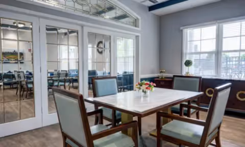 Small dining room with a square table and four chairs, mirrored French doors, and a bright window.