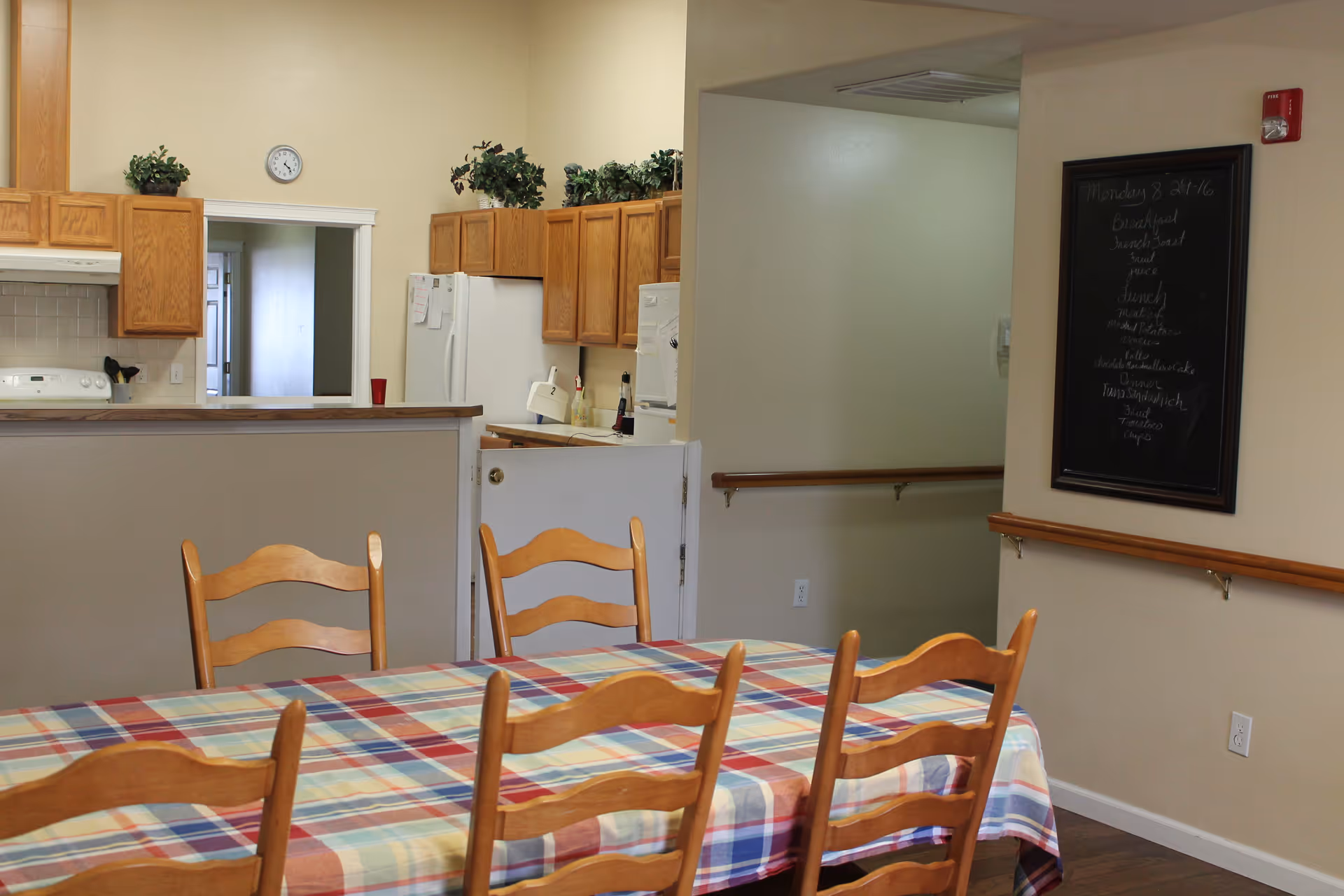 Dining area with a wooden table covered by a colorful plaid tablecloth and six wooden chairs. In the background, there is a kitchen with wooden cabinets, a white refrigerator, and a stove. A small clock is mounted on the wall above a pass-through window. A blackboard with handwritten text is mounted on the wall to the right.