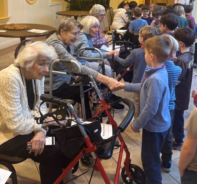 Several elderly women seated in a row, some using walkers, are interacting and shaking hands with a group of young children standing in front of them in a communal indoor space.