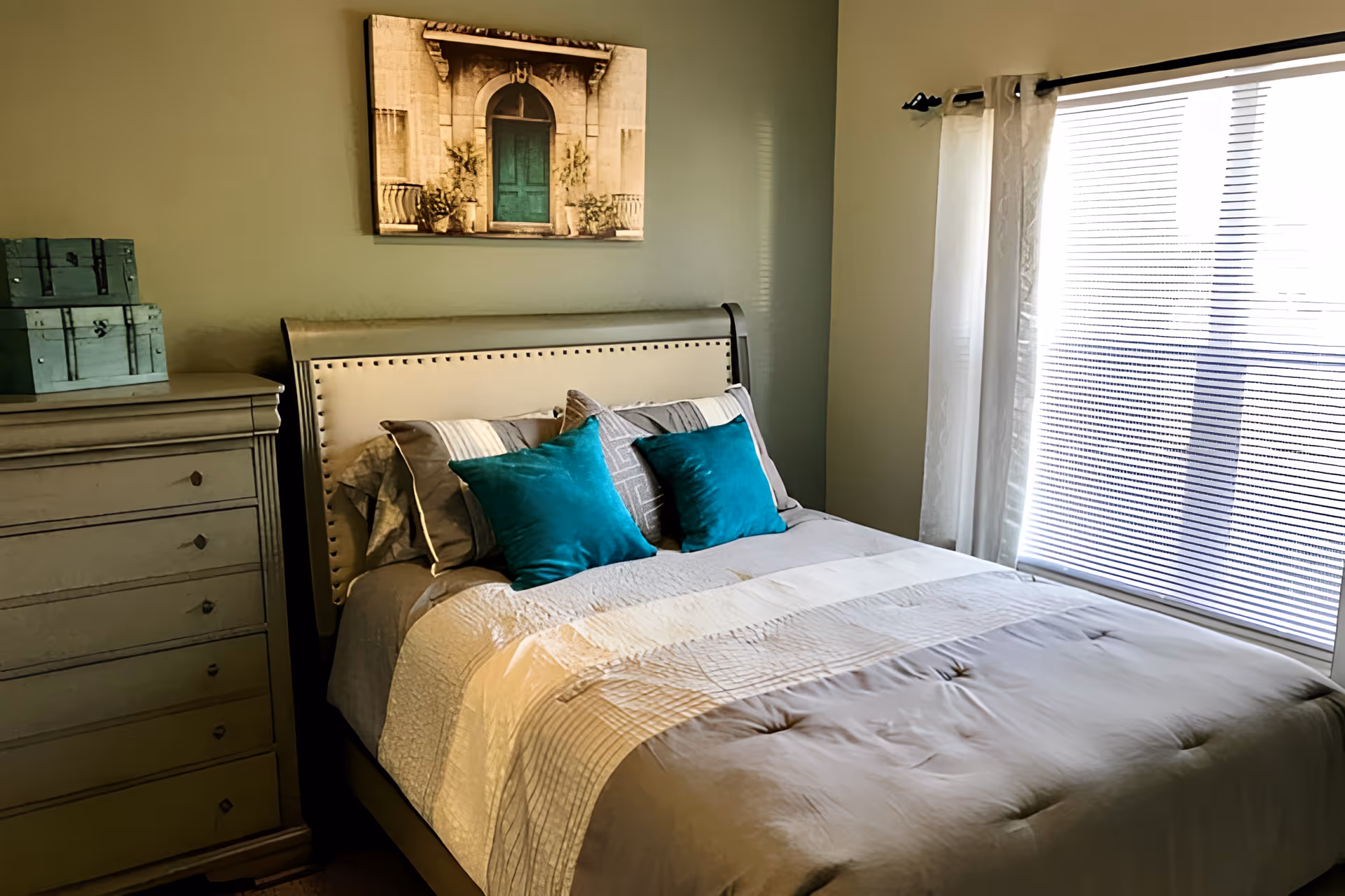 A cozy bedroom with a neatly made bed featuring gray and white bedding and two teal accent pillows. To the left of the bed is a gray wooden dresser with two teal decorative boxes on top. Above the bed hangs a framed picture of a green door with an archway. On the right side, there is a large window with white curtains and blinds partially closed, letting in natural light.