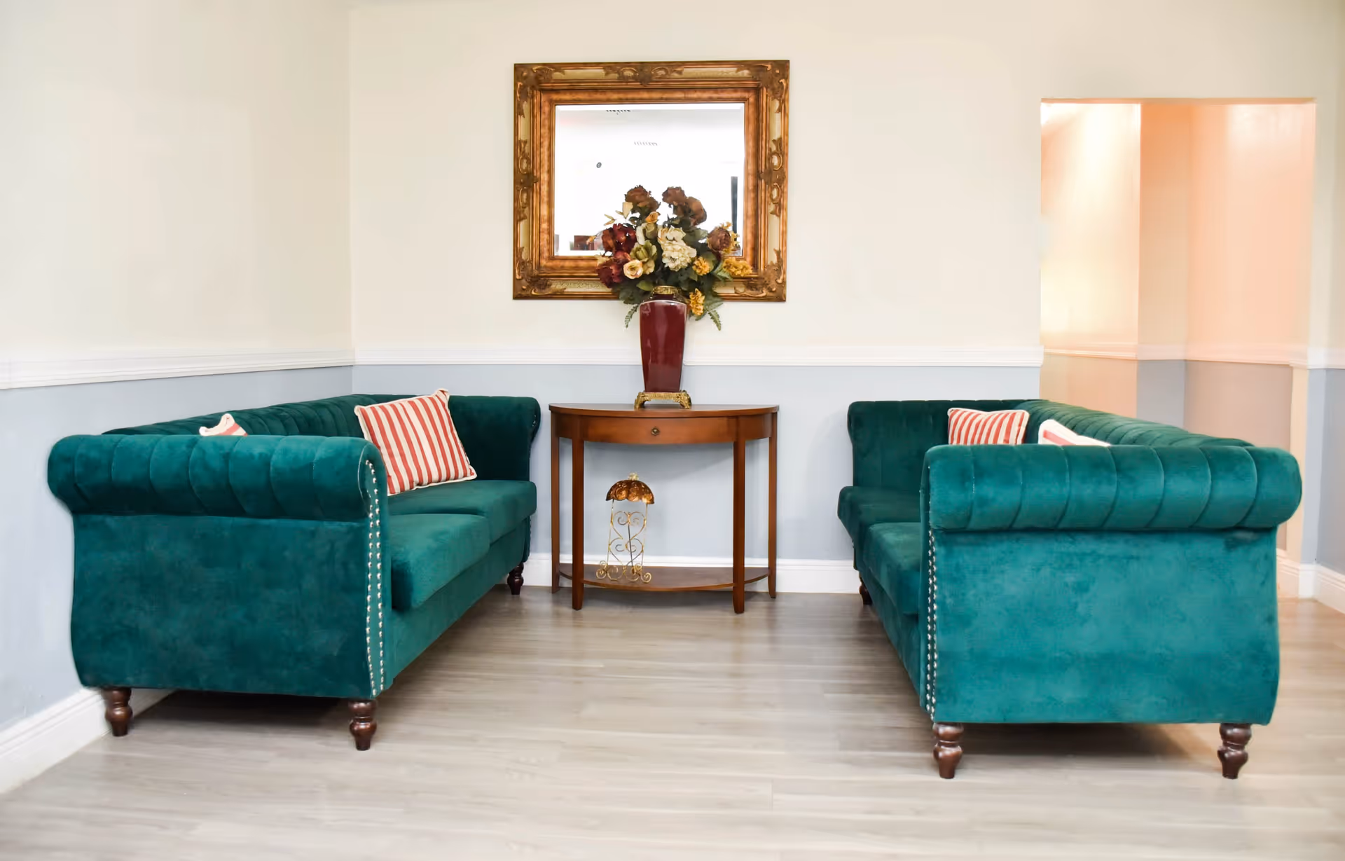 A cozy seating area with two green velvet sofas facing each other, each with striped red and white pillows. Between the sofas is a wooden console table with a red vase holding a bouquet of flowers. Above the table hangs a large ornate gold-framed mirror. The room has light-colored walls with a white chair rail and light wood flooring.