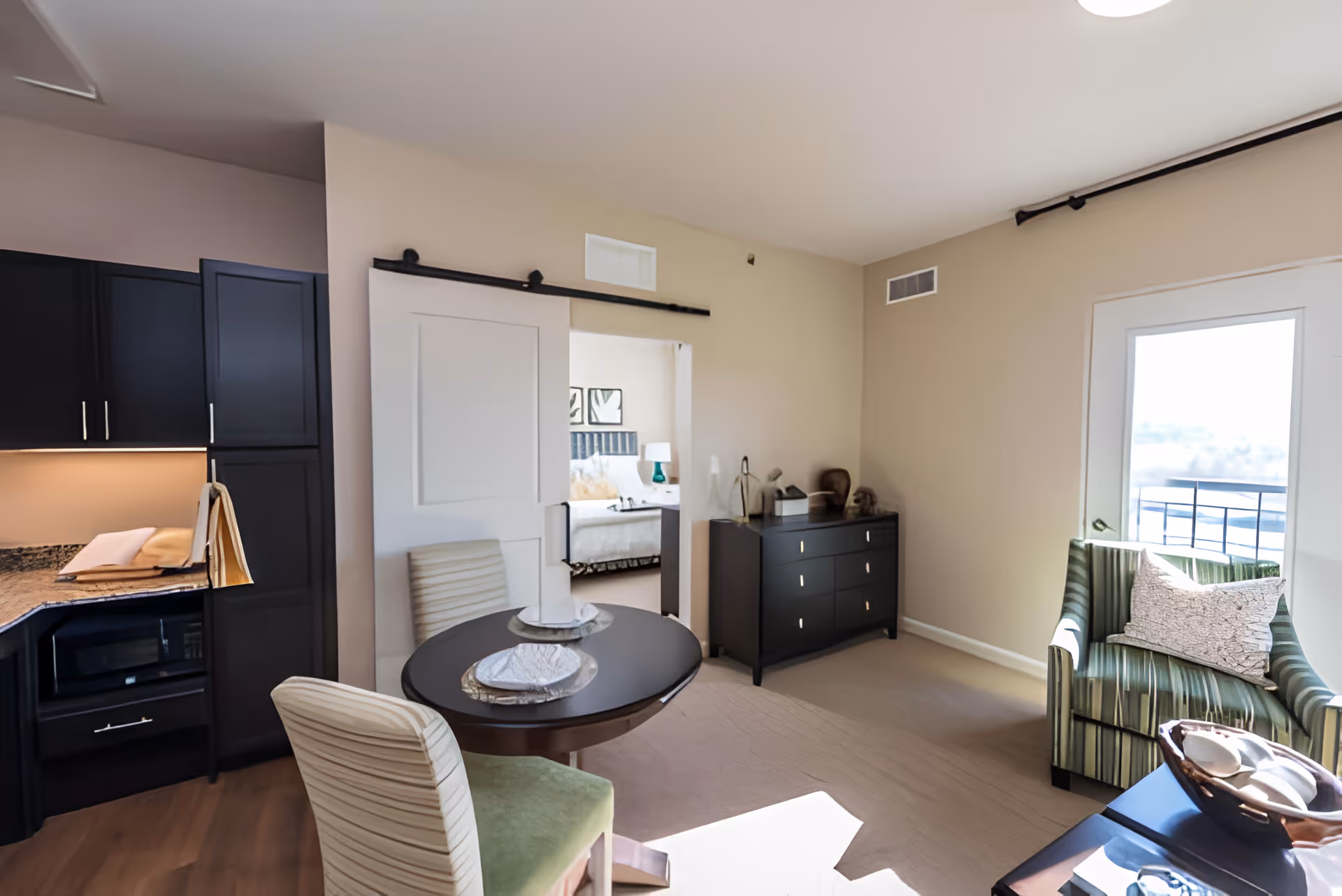 A bright living area in a senior living facility featuring a small round dining table set for two with striped chairs, a green striped armchair with a decorative pillow near a glass door leading outside, a black dresser with decorative items, and a partial view of a kitchen with dark cabinets. A sliding barn door opens to a bedroom with a bed and bedside lamp visible.