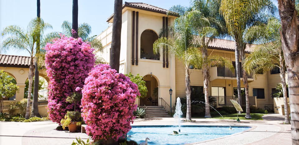 Outdoor courtyard area of a senior living facility with a central water fountain surrounded by palm trees and large bushes with vibrant pink flowers. The building in the background has a beige exterior with arched doorways and balconies.