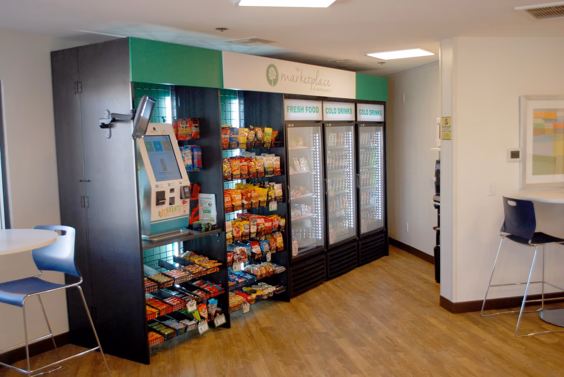 A small convenience market area inside a facility with shelves stocked with snacks and three refrigerated glass-door coolers labeled Fresh Food and Cold Drinks. There is a self-checkout kiosk on the left side and seating with tables and chairs nearby. The floor is wooden and the walls are light-colored with framed artwork.