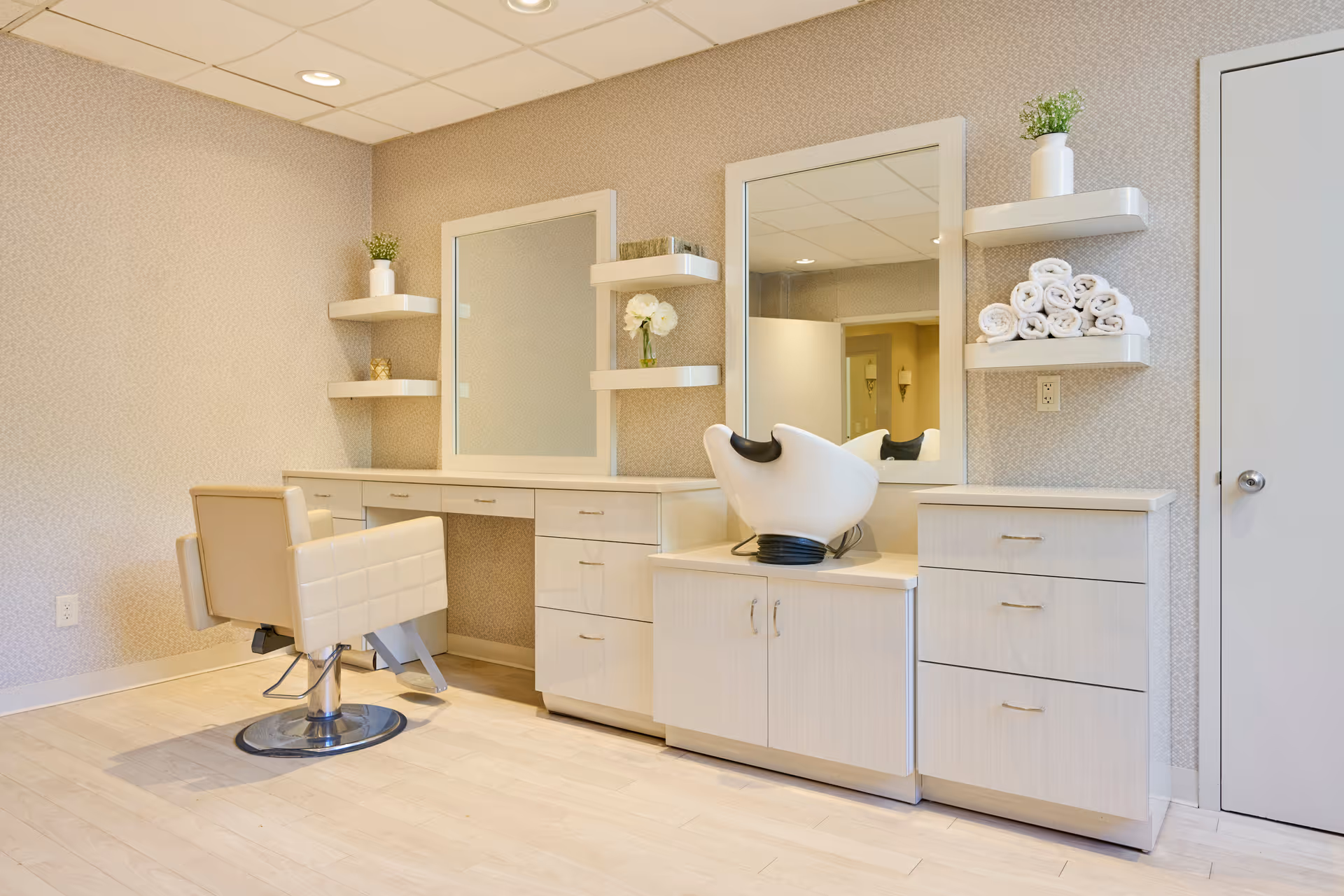 A clean and modern salon area in a rehabilitation facility featuring a beige salon chair, two large mirrors mounted on the wall, white cabinetry with drawers and shelves, neatly rolled white towels on a shelf, and small potted plants and flowers for decoration.
