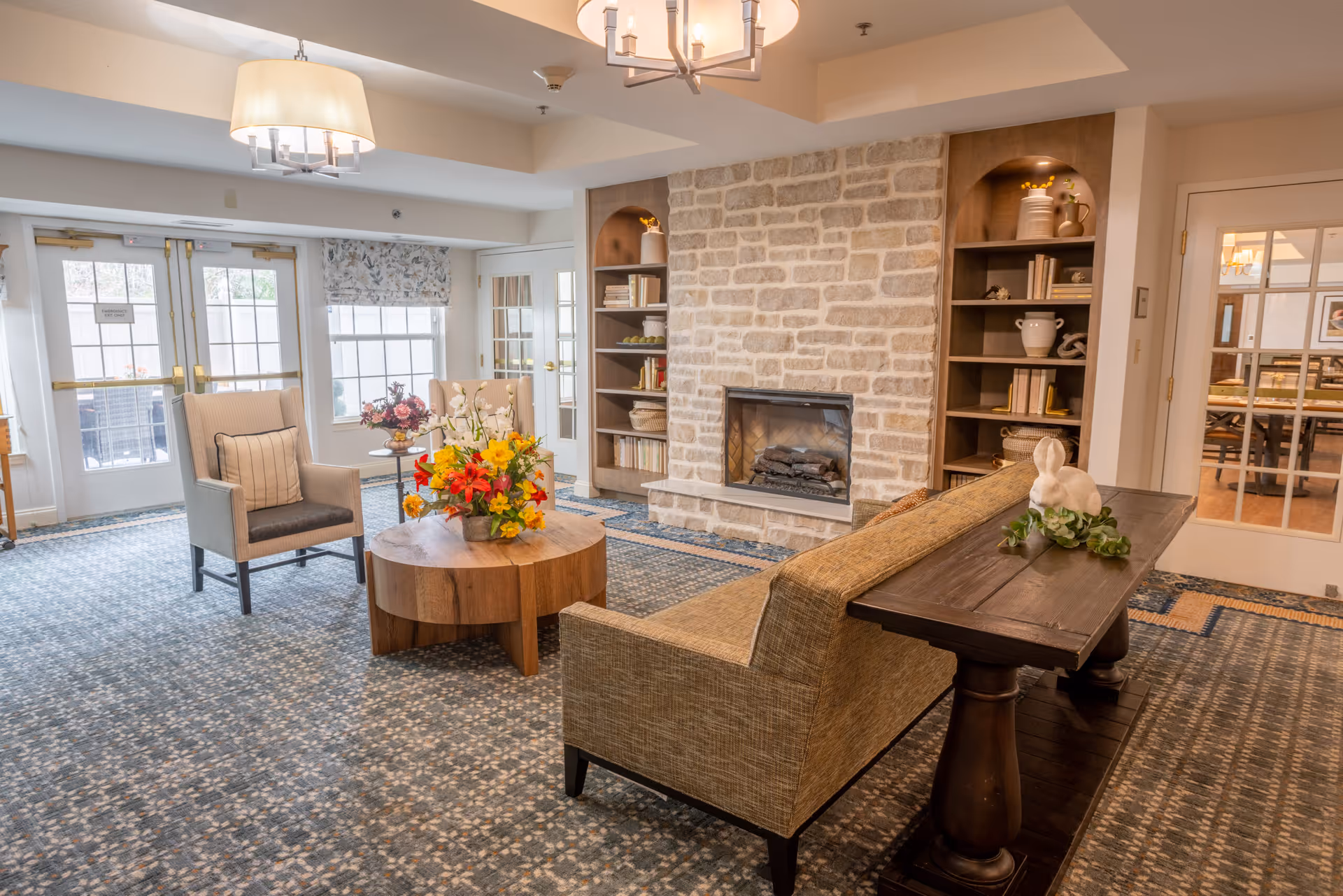 A cozy living room area in a senior living facility featuring a stone fireplace flanked by built-in shelves with decorative items and books. The room has a patterned carpet, a wooden coffee table with a colorful flower arrangement, two armchairs, and a sofa with a wooden console table behind it. Large windows and glass doors allow natural light to enter the space.