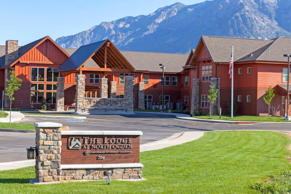Exterior view of The Lodge at North Ogden, a large red building with stone pillars and a covered entrance, set against a backdrop of mountains under a clear blue sky. A stone and wood sign with the facility's name is visible in the foreground on a well-maintained lawn.