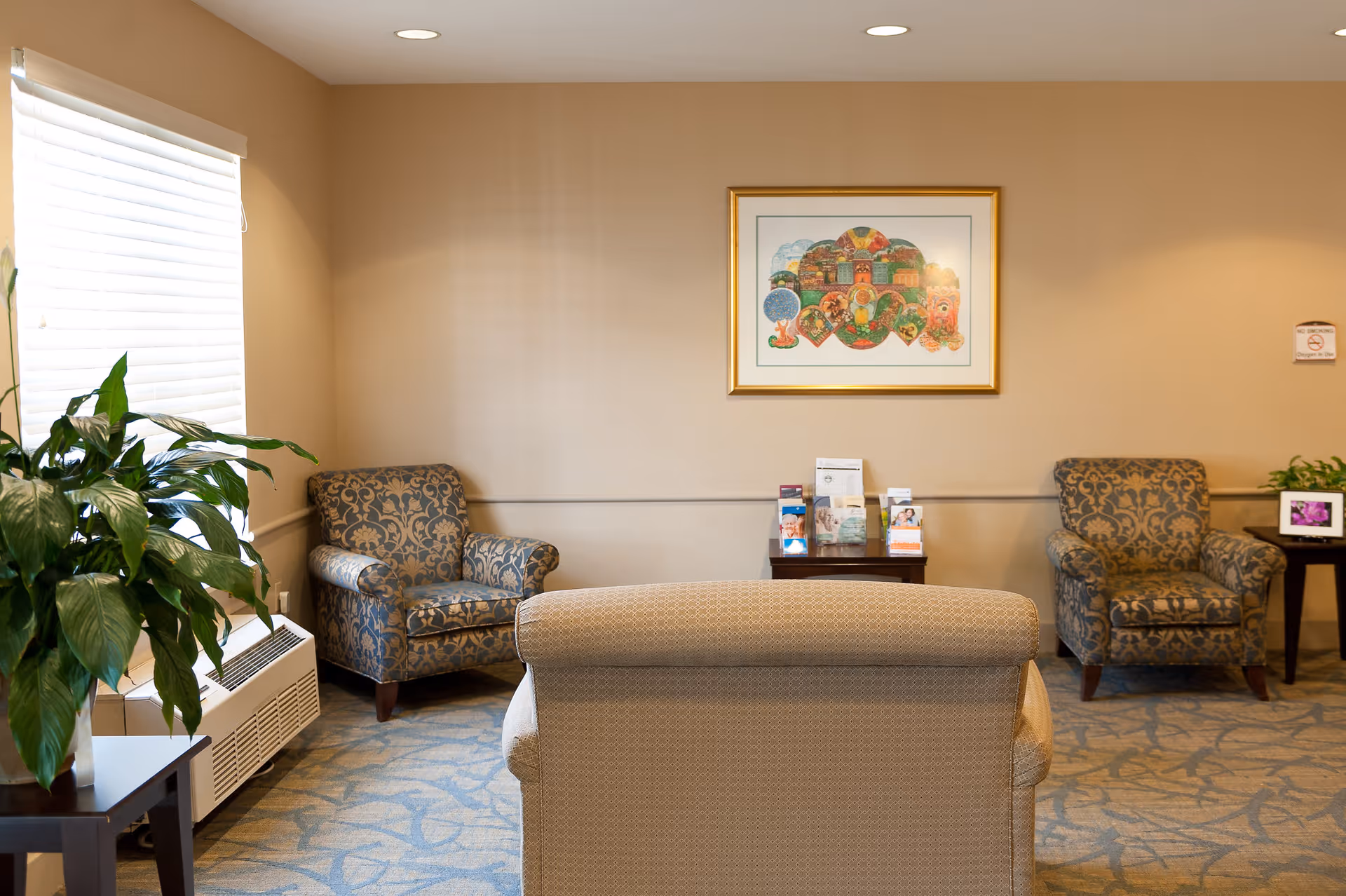 A cozy sitting area in a nursing and rehabilitation center featuring three upholstered armchairs arranged around a small wooden table with brochures. The walls are beige with a framed colorful artwork hanging above the table. There is a large green plant on a side table near a window with white blinds, and the floor is carpeted with a blue and beige patterned rug.