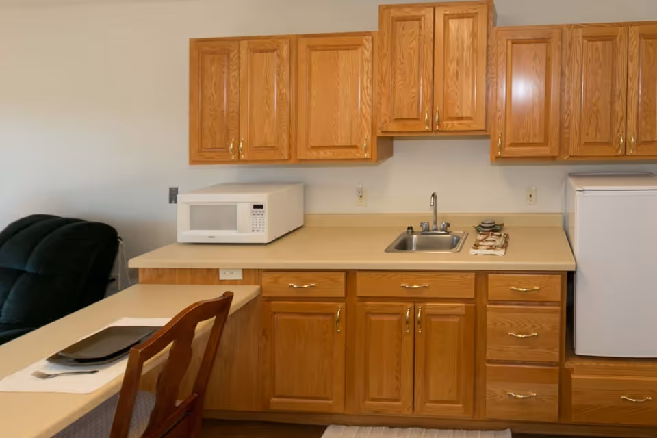 A kitchen area with wooden cabinets, a countertop with a sink, a white microwave, and a small refrigerator. A wooden chair is positioned at the counter with a placemat, plate, and utensils. In the background, part of a dark green recliner chair is visible.