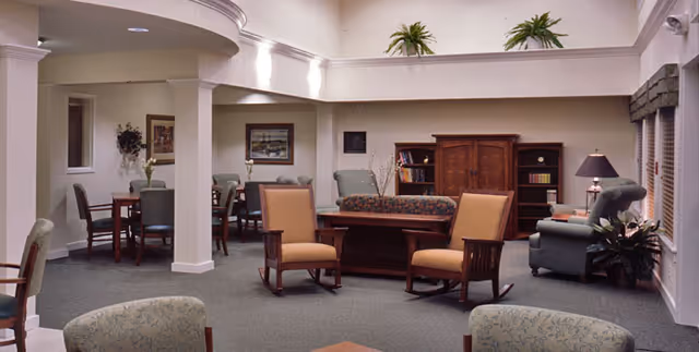 A spacious living room area in a senior living facility with multiple seating options including armchairs and sofas arranged around a central wooden coffee table. The room features high ceilings with recessed lighting, decorative plants on a ledge above, framed artwork on the walls, and large windows with blinds allowing natural light to enter.