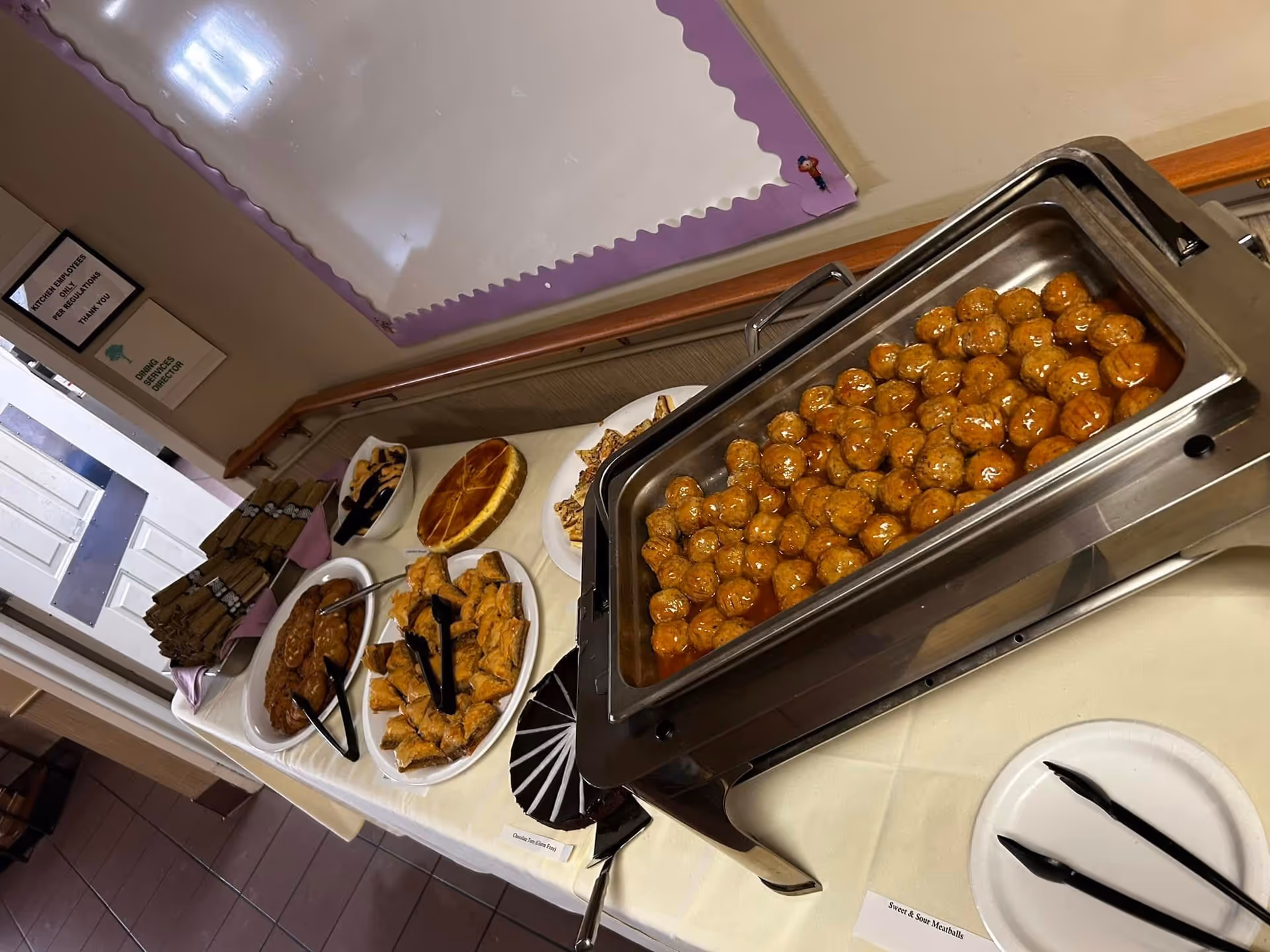 A buffet table set up with various food items including a tray of sweet and sour meatballs, plates of pastries, a pie, and wrapped utensils. The table is covered with a white tablecloth and is located indoors near a door and a bulletin board with a purple border.