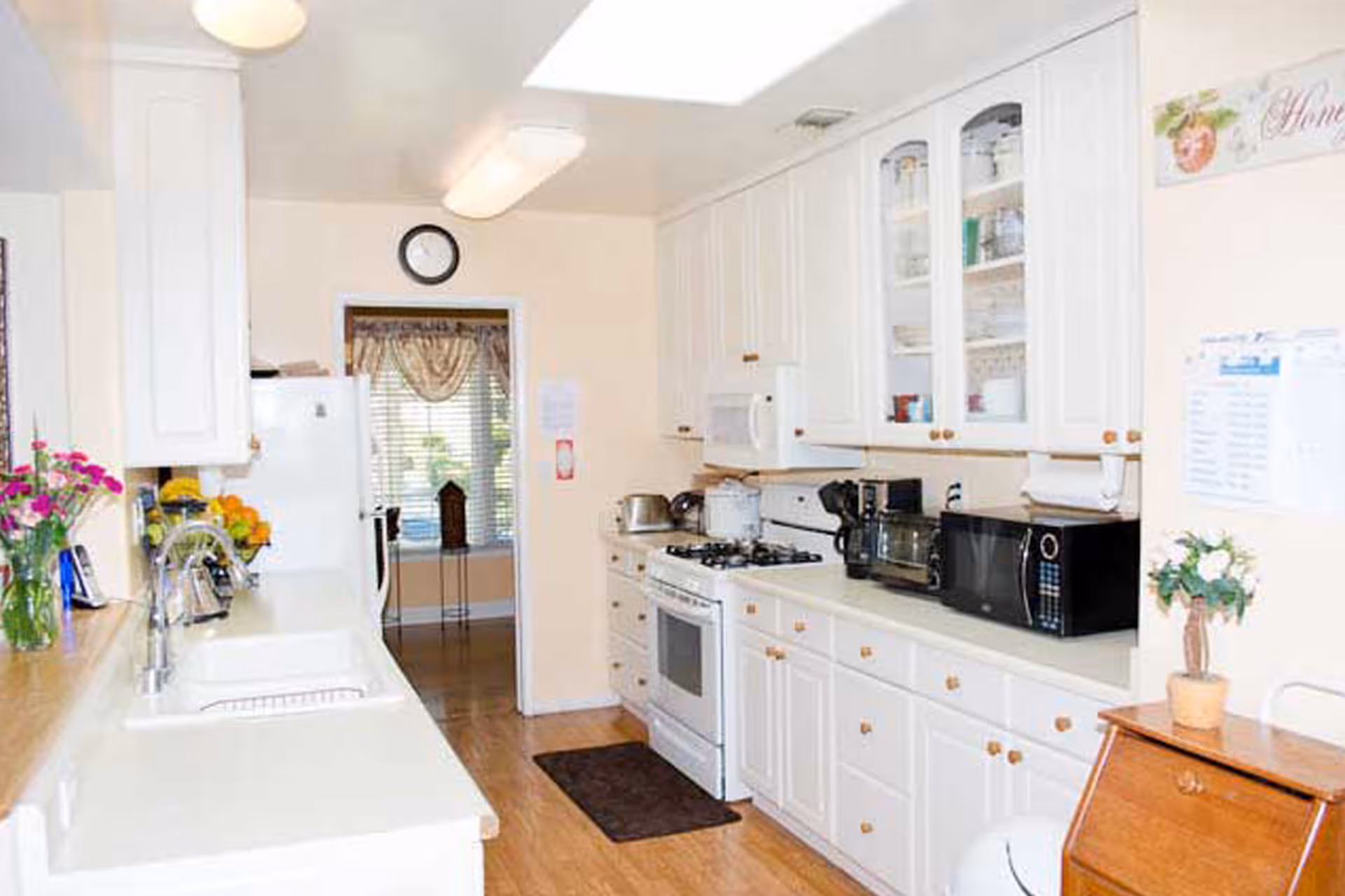 Bright galley-style kitchen with white cabinets, countertops, a stove and microwave, and flowers on the counter.