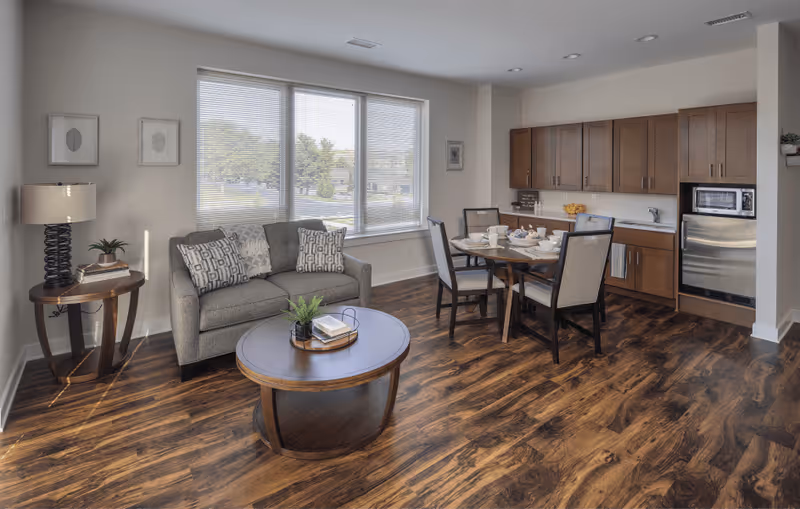 A bright and cozy living area with a gray loveseat adorned with patterned pillows, a round wooden coffee table with a small plant and books, a wooden side table with a lamp and plant, and a dining area with a round table set for four. The background features large windows with blinds and a kitchenette with wooden cabinets, a microwave, and a small refrigerator.