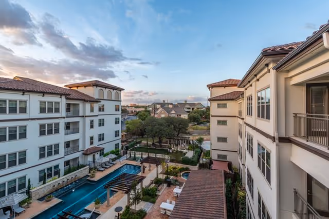View of an outdoor courtyard area at Overture Stone Oak featuring a swimming pool, lounge chairs, trees, and surrounding multi-story residential buildings under a partly cloudy sky at sunset.