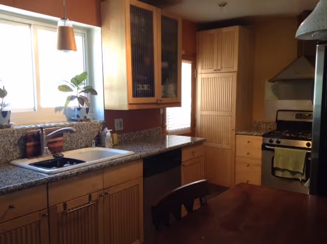 A kitchen interior with wooden cabinets, granite countertops, a double sink under a window with potted plants, a dishwasher, a stainless steel stove with a yellow towel hanging on the handle, and a wooden dining table with chairs.