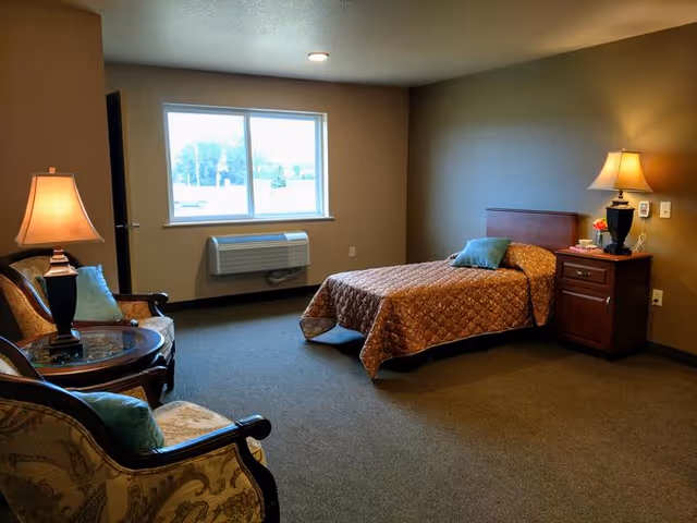A simple, cozy bedroom in an assisted living facility featuring a single bed with a brown quilt and a blue pillow, a wooden nightstand with a lamp and a small flower vase, two upholstered armchairs with a round glass-top table between them, and a window letting in natural light.