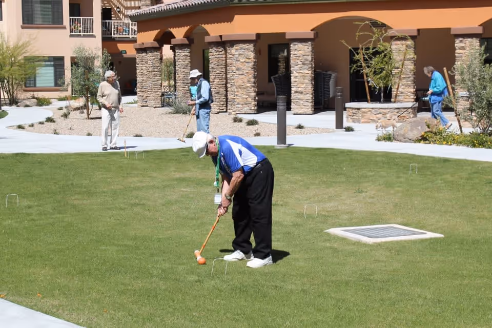 Four elderly people playing croquet on a green lawn outside a building with stone pillars and tan walls. One man in the foreground is preparing to hit a ball with a mallet, while others stand or walk nearby, including a person walking a small dog.
