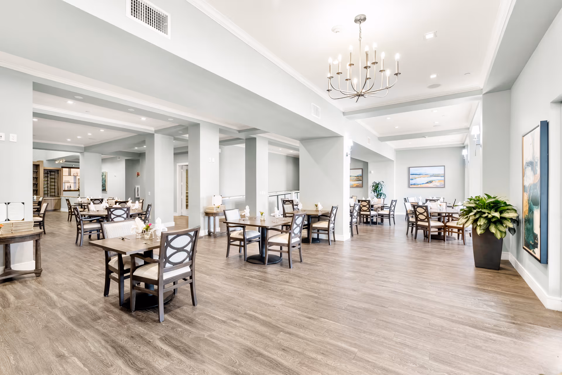 A spacious and bright dining room with multiple wooden tables and chairs arranged neatly. The room features light gray walls, wood flooring, modern chandeliers, and framed artwork on the walls. There are also some potted plants adding greenery to the space.