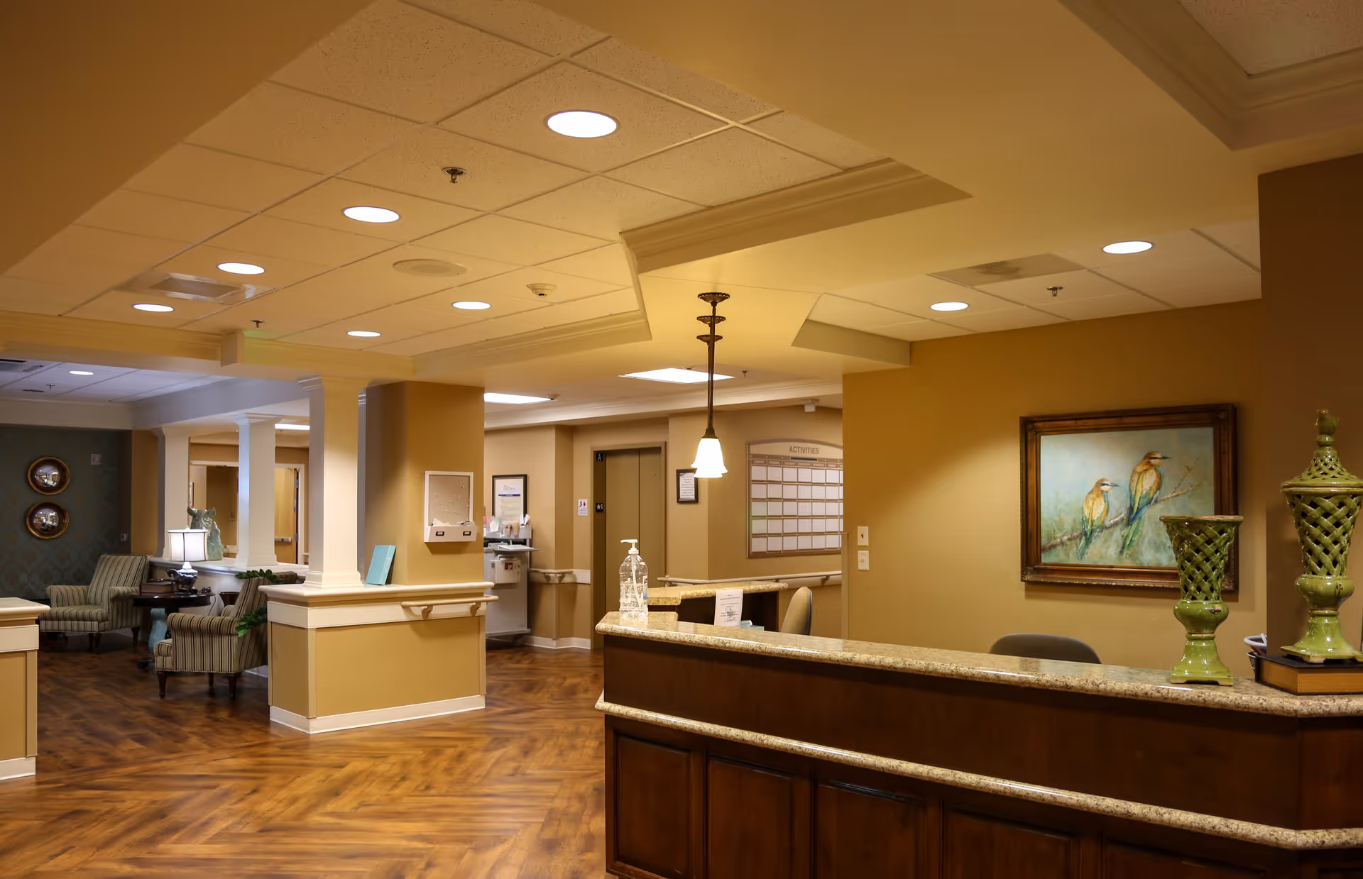 Interior view of a senior living facility reception area with a wooden front desk, decorative green vases, a painting of two birds on the wall, and a seating area with armchairs and a lamp in the background. The space has warm lighting and wood-patterned flooring.