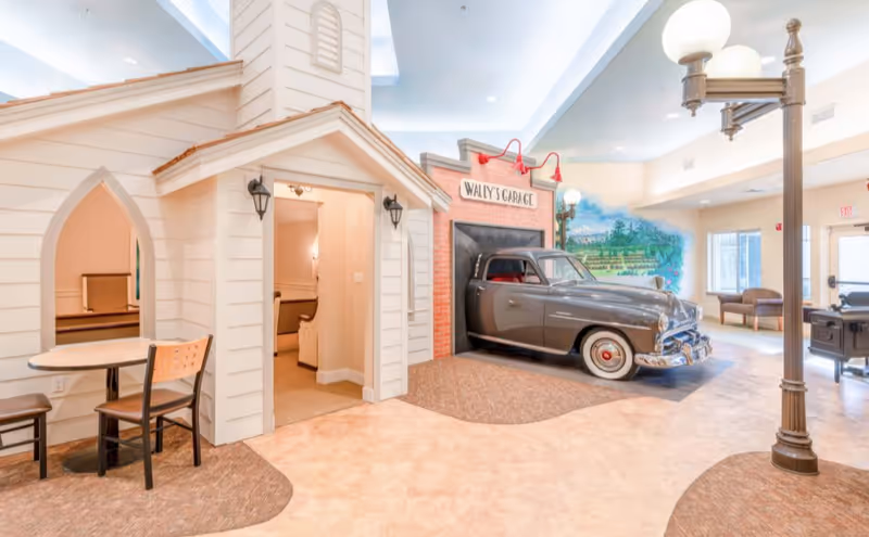 Interior of a senior living facility featuring a playful setup with a small house structure, a table with chairs, and a vintage car parked in front of a faux garage labeled 'Wally's Garage'. The room is well-lit with large windows, a mural on the wall, and a streetlamp-style light fixture.