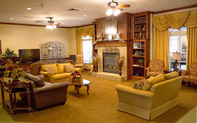A cozy living room area with multiple sofas and armchairs arranged around a wooden coffee table. There is a fireplace with decorative vases and a framed picture above it. Built-in wooden shelves flank the fireplace, holding books and decor. Large windows with yellow curtains allow natural light into the room. A television and a large birdcage are visible in the background.