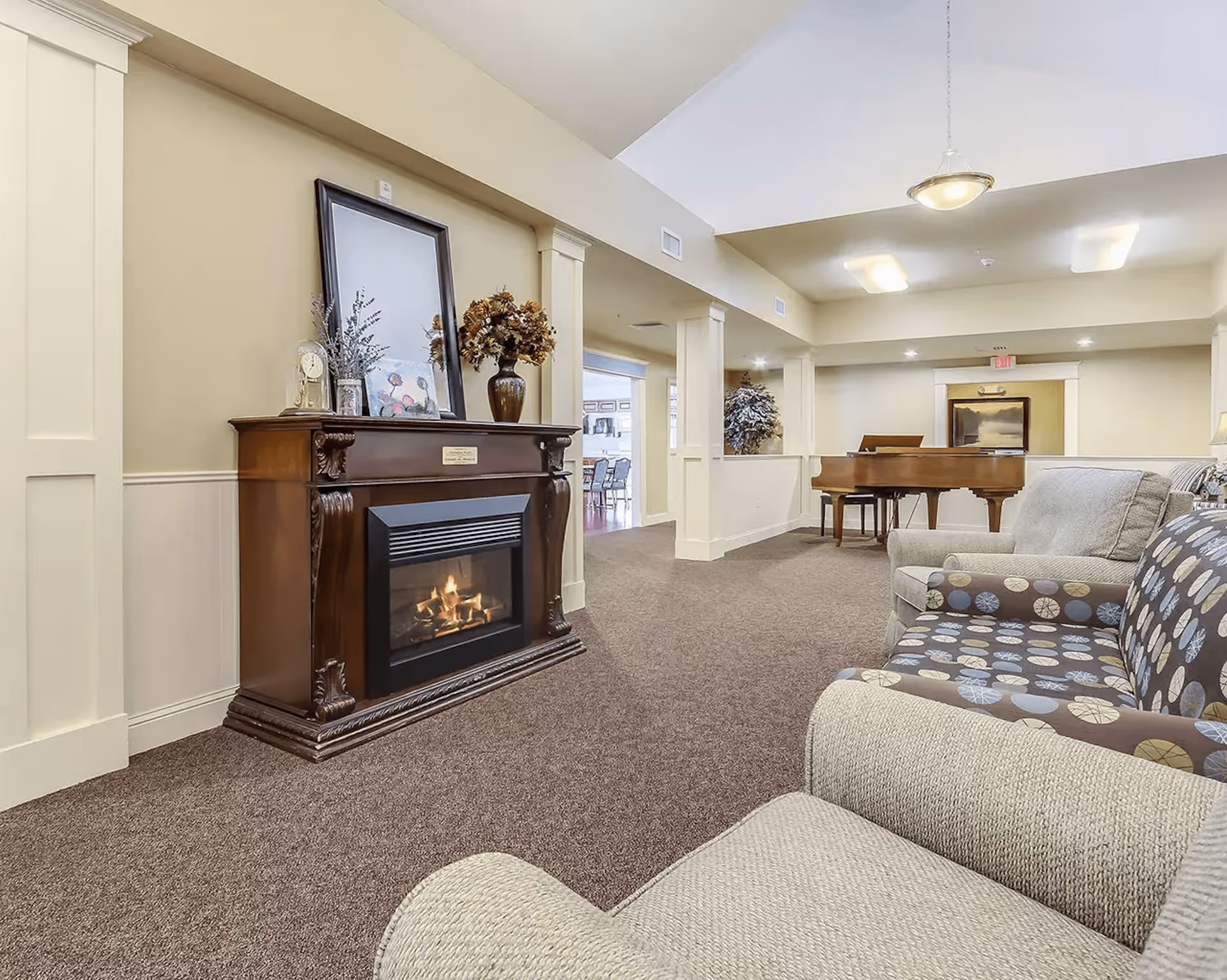 A cozy assisted living facility common area featuring a lit electric fireplace with decorative items on the mantel, comfortable upholstered chairs with patterned and solid fabric, and a grand piano in the background under soft ceiling lighting.